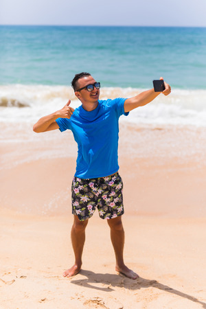 Man in sunglasses sits with a smartphone in the hands and making selfie on the phone near the sea. Summer timeの写真素材