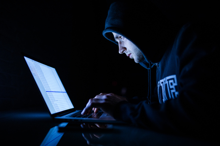 Man in a hood working in front of a laptop hacker cracking programs in the night. Hacker
