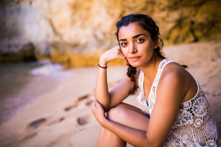 Potrait latin girl sitting on golden sand at beach near rocks and looking at ocean.の写真素材