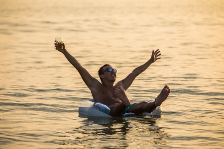 Young man with raised hands in the ocean or sea water with glass of beer screaming and enjoy summer vocation on on inflatable ring on sunset.の写真素材