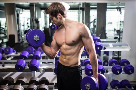 Athletic shirtless young male fitness model holds the dumbbell with light isolated on dark background.の写真素材