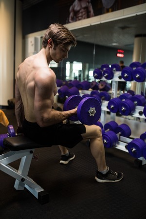 Athletic shirtless young male fitness model holds the dumbbell with light isolated on dark background.の写真素材