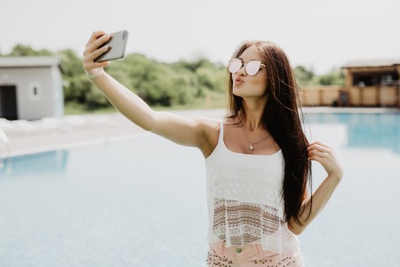 Close-up selfie-portrait of attractive brunette girl with long hair standing near pool. She wears pink T-shirt, sunglasses. She is making a kiss to the camera.の写真素材