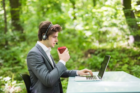 Side portrait Businessman with headset sitting at the office desk with laptop computer and cup of coffee in call center in green forest park.の写真素材