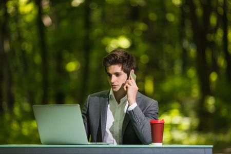 Portrait of Businessman sitting at the office desk work at laptop computer and cup of coffee talking mobile phone on road of green forest park. Business concept.の写真素材