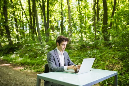Portrait of Businessman sitting at the office desk work at laptop computer in green forest park. Freelancer working on office table in green park.の写真素材