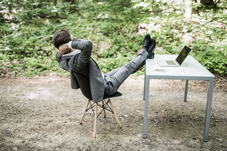 Relaxed business man in suit with heads over head and legs on office desk after successful work in green park. Freelancer relax after great deal in green forest.の写真素材