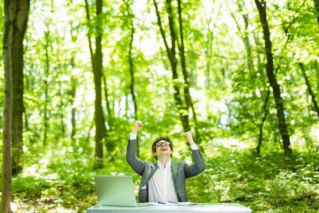 Portrait of handsome succesful business man in suit working at laptop at office table with raised hands in green forest park.の写真素材