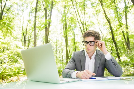 Portrait of handsome business man in suit working at laptop at office table thinking in green forest park. Business concept.の写真素材
