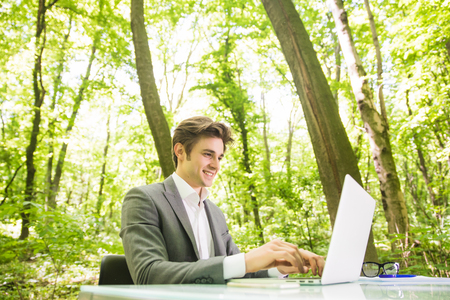 Portrait of young handsome business man in suit working at laptop at office table in green forest park. Business concept.の写真素材