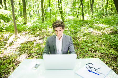 Portrait of young handsome business man in suit working at laptop at office table in green forest park.の写真素材