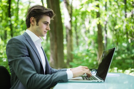 Portrait of handsome business man in suit at laptop at office table in green forest park. Business concept.の写真素材