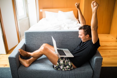 Man get good news from laptop with happy victory emotion. Side view of handsome young man using his laptop with smile while sitting on the couch at homeの写真素材