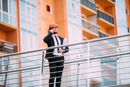 Engineer with plan in his hand talking on cell phone on construction building side.
