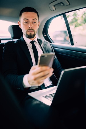 Confident young businessman working on his laptop and talking on the phone while sitting in the carの写真素材