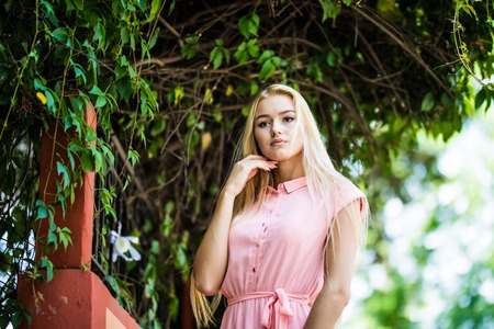 Beautiful blonde woman in green leaves background. Young girl wearing dress standing in the street. Pretty female with frizzy hairstyle and blue eyes.の写真素材