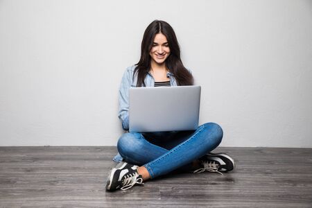 Young woman sitting on the floor with crossed legs and using laptop on gray backgroundの写真素材