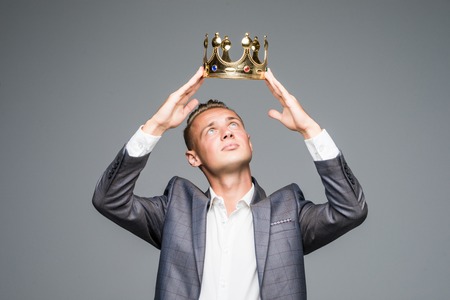 Young attractive man in suit holding above his head a golden crown on a gray backgroundの写真素材