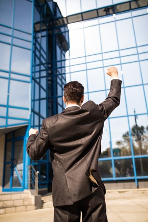 Celebrating success. Back view of excited young businessman with raised arms expressing positivity while standing outdoors with office buildingの写真素材