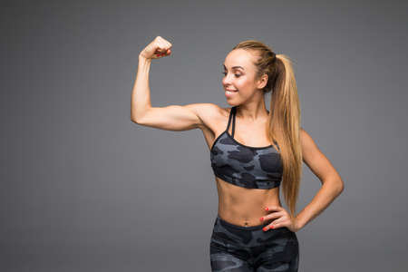 Cheerfully smiling mixed race sporty woman demonstrating biceps, isolated on grayの写真素材