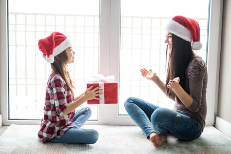 Young happy mother with her daughter holding teddy bear while sitting on white carpet near Christmas treeの写真素材