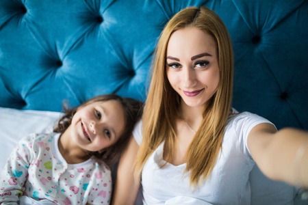 Happy mother and daughter taking selfie with smartphone on bedの写真素材