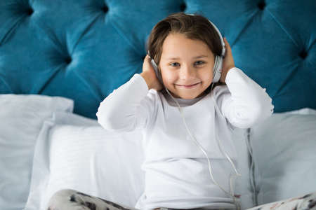 Cheerful girl listening to music and dancing while sitting in her bed, looking at cameraの写真素材