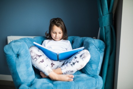 Funny happy toddler girl reading a book and playing in bed. Kids play at home. White nursery. Child in sunny bedroom. Children read and study. Interior for baby and young kid.の写真素材