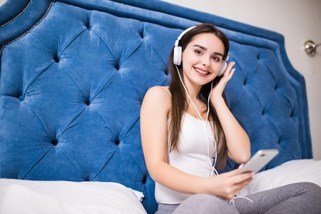 Smiling lady in pajamas listening music with smartphone and headphones while sitting in bedの写真素材