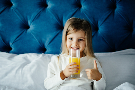 Beautiful girl kid holding glass of orange juice, showing thumb up gesture, looking at camera while sitting in bedの写真素材