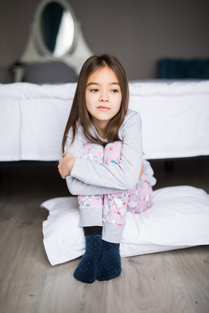 Portrait of an upset little girl sitting on the floor near bed and looking at camera in the morningの写真素材