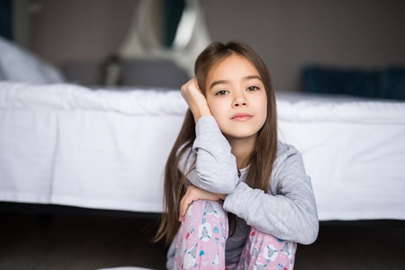 Portrait of an upset little girl sitting on the floor near bed and looking at camera in the morningの写真素材