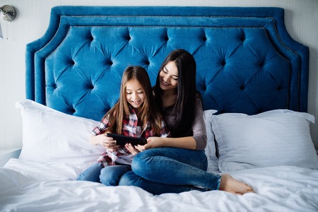 Beautiful young woman and her charming little daughter are using a digital tablet and smiling while...