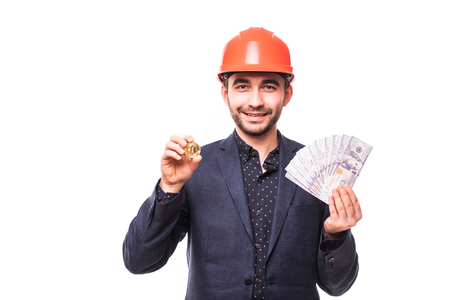 Handsome man miner in protective hemlet with bitcoin in one hand and cash dollars in other isolated on white backgroundの写真素材