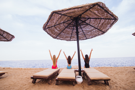 Back view portrait of three women friends sitting and resting in lounge chairs with umbrella on the sea beach.の写真素材