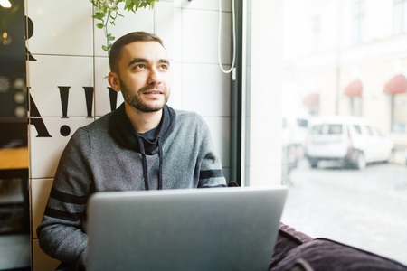 Handsome male ur typing message to content manager of website of marketing corporation on keyboard of laptop computer arranging meeting to discuss collaboration while sitting in cafe.の写真素材