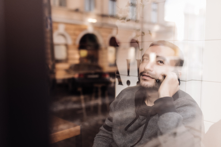 Enjoying his phone call. Handsome man talking on mobile phone and looking throughwindow with smile while sitting in cafeの写真素材