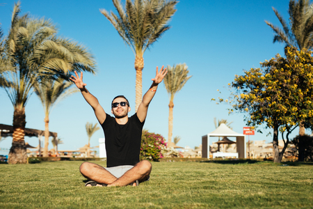 Handsome bearded man in sunglasses sitting on green grass and relax enjoy summer vocation with raised hands up on palms and blue sky background.の写真素材