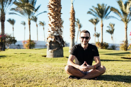 Handsome bearded man in sunglasses sitting on the grass under palms on summer vocation luxary resort