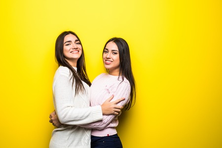 Portrait of two happy girls dressed in sweaters hugging over yellow background