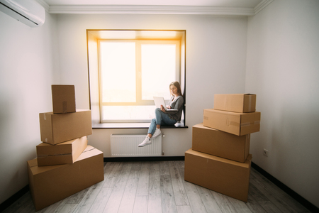 Attractive young woman is moving, sitting among cardboard boxes, using a laptop and smilingの写真素材