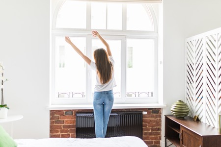 Woman Stretching in the Bedroom at the Window after Wake up. Rear view, Model in the Bedroom looks out the Window with his Arms Raised over his head. Girl in the Morning Pajamasの写真素材