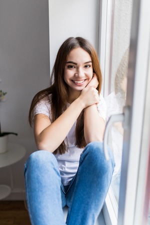 Relaxing. Attractive young woman smiling and wearing a shirt and jeans and sitting on the windowsillの写真素材