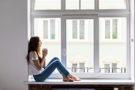 Beautiful daydreamer. Beautiful young woman holding coffee cup while sitting at windowsill at homeの写真素材