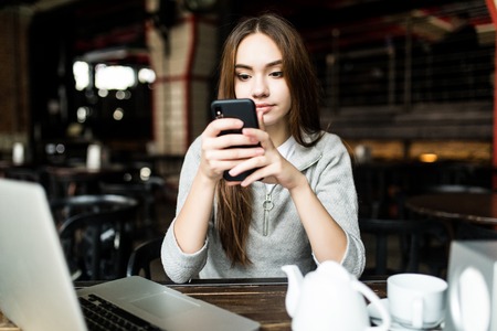 Young woman or successful working on smart phone and laptop computer at coffee shop interior. Girl student sitting in university cafeteria while using technology, internet distance workの写真素材