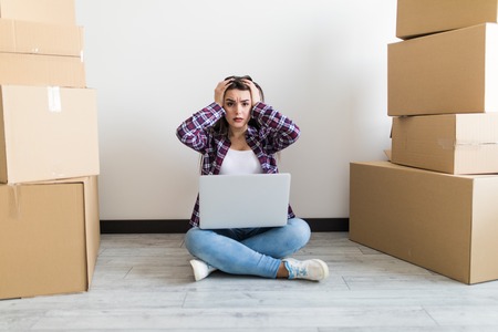 Beautiful woman moving in her house and unpacking, she is sitting on the floor surrounded by boxes, using a laptop looking shocked on screenの写真素材