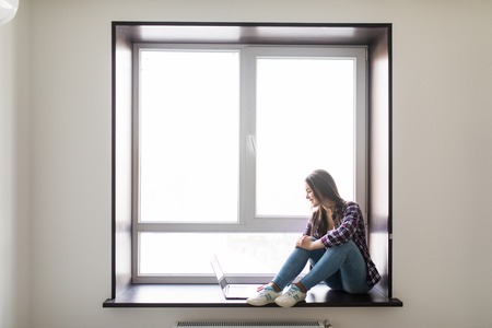Beautiful young woman sitting on windowsill and using laptopの写真素材