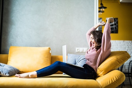 Woman raising her hands up on the couch with laptop at home