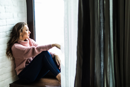Smiling pretty woman in sweater while sitting on a windowsill at homeの写真素材