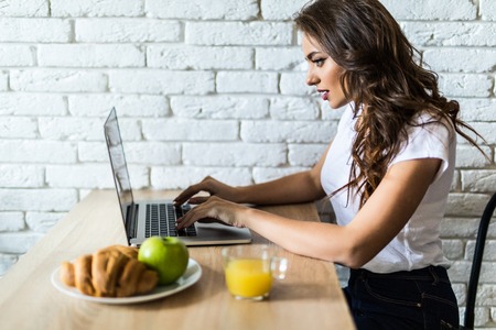 Young cheerful woman using on laptop computer and eating fruits in the kitchenの写真素材